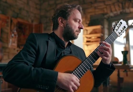 Marcin Dylla podczas dynamicznego występu na scenie, grający na gitarze klasycznej, oświetlony ciepłym światłem scenicznym.