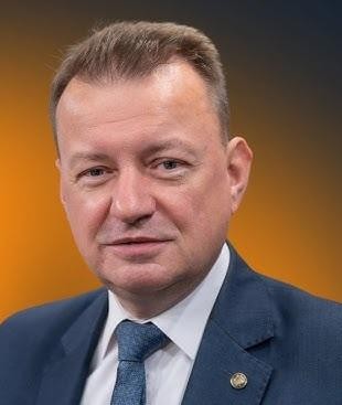 Mariusz Błaszczak podczas inspekcji sprzętu wojskowego lub wizyty na poligonie, symbolizującej jego rolę w ministerstwie obrony.