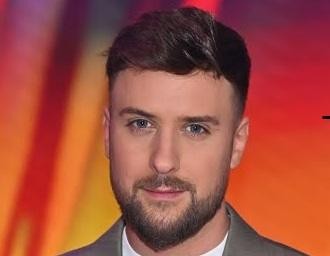Aleksander Sikora, prezenter telewizyjny, uśmiecha się subtelnie, stojąc za kulisami lub w profesjonalnym otoczeniu.
