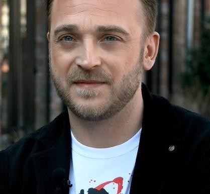 Mateusz Gessler jako juror programu kulinarnego, uśmiechnięty, w tle studio telewizyjne z elementami kuchni.
