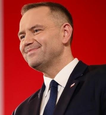 Karol Nawrocki przemawiający na konferencji historycznej, z gestykulacją podkreślającą jego zaangażowanie, na tle ekranu z historycznymi grafikami.