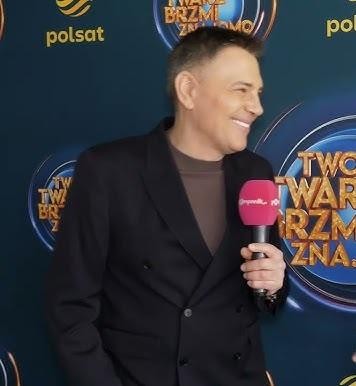 Krzysztof Ibisz w akcji na planie telewizyjnym, z uśmiechem prezentujący program.
